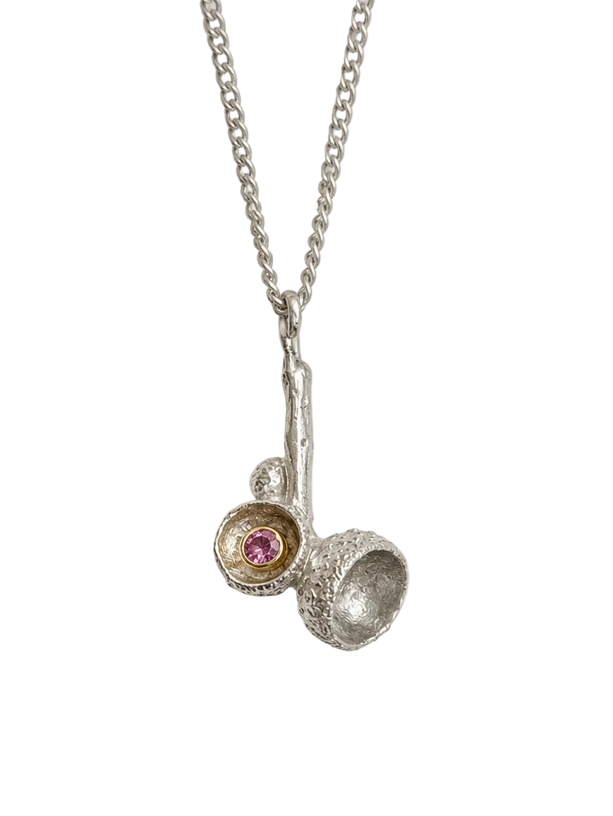 A silver pendant necklace featuring a textured silver branch-like charm with two small cup-like settings, one holding a tiny pink gemstone and the other textured, hanging from a delicate silver chain against a light gray background.