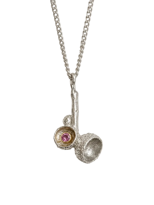 A silver pendant necklace featuring a textured silver branch-like charm with two small cup-like settings, one holding a tiny pink gemstone and the other textured, hanging from a delicate silver chain against a light gray background.